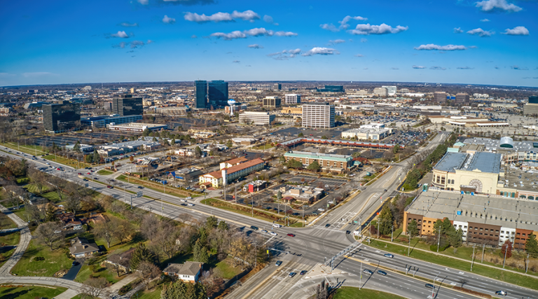 Aerial view of Schaumburg, Illinois showing busy streets, office buildings, and shopping areas under a clear blue sky.